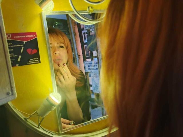 PaulettWatsoon with red hair, looking at herself in a yellow mirror, pouting with fingers on her lips, reflection shows her in a green shirt, mirror has a light.
