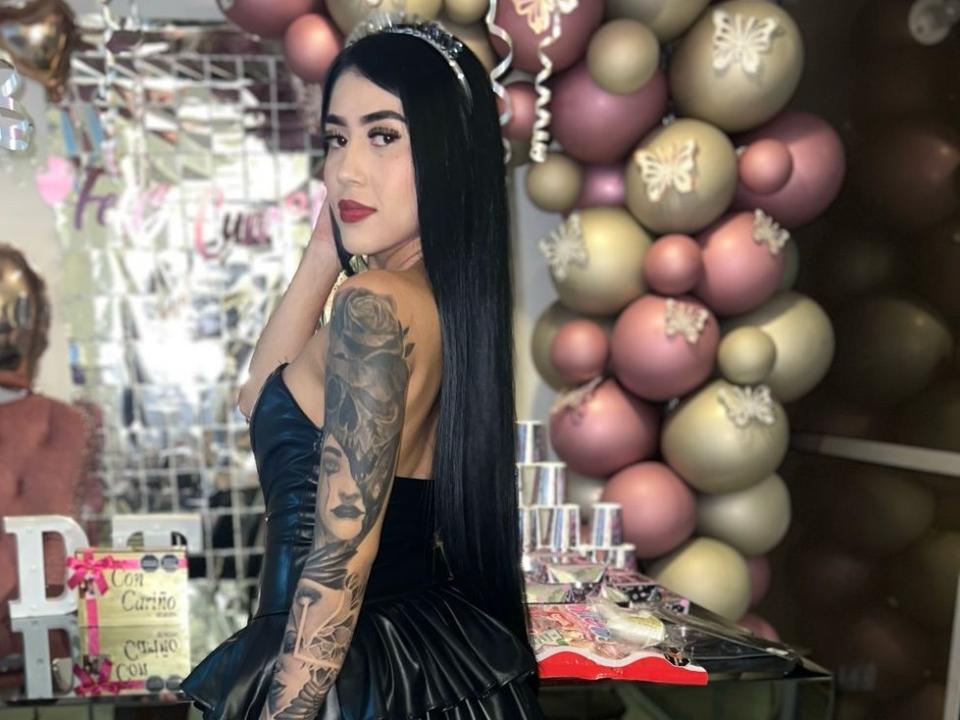 LauraSantorini with long black hair, red lipstick, and a black strapless dress, showing her tattooed arm and looking over her shoulder. Pink and gold balloons in the background.