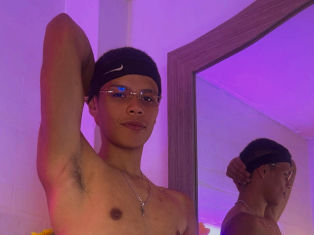 Shirtless JordanKing with a black Nike beanie, glasses, and necklace, arms raised, showing armpit hair, reflected in a mirror with purple lighting.