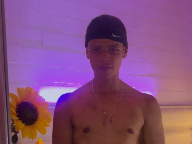 Shirtless JordanKing with dark skin, black Nike cap, glasses, necklace, and a sunflower to the left. Purple and yellow light casts a glow on his upper body.