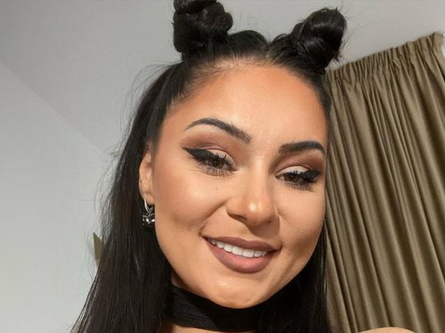 PrettyA has black double buns, thick eyeliner, long lashes, and a smile showing her teeth. She's wearing a black choker and has tan skin with dark brown hair.