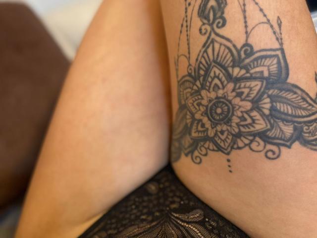 A coxa de MilaDor, com uma tatuagem detalhada em preto em formato de mandala em um tom de pele claro, sentada com o bumbum em uma roupa íntima de renda preta, com foco na tatuagem e na renda..