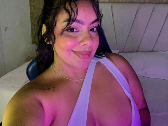 AmarantaCarey with dark wavy hair, pink nose ring, and white deep cleavage dress, smiling at camera, showing large tits and small tattoo on left shoulder. Bedroom setting.