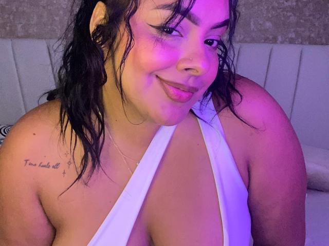 AmarantaCarey with wet black hair, white halter top, showing large boobs, tattoo on left shoulder, nose ring, smiling at camera, purple lighting, slightly messy makeup, confident expression.