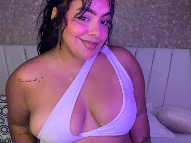AmarantaCarey with large tits in a white, deep-cut halter top, smiling with purple lighting on her tan skin, black hair, and tattoo on her right shoulder.