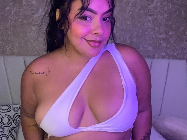 AmarantaCarey with large, rounded boobs in a white, deep-cut halter top, smiling at the camera, has wavy black hair, and tattoos on her right shoulder and left arm.