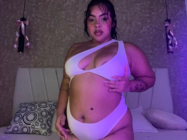 AmarantaCarey with dark wet hair, wearing a white cutout bikini that exposes her large tits and a small black mole on her belly. She has tattoos on her right arm.
