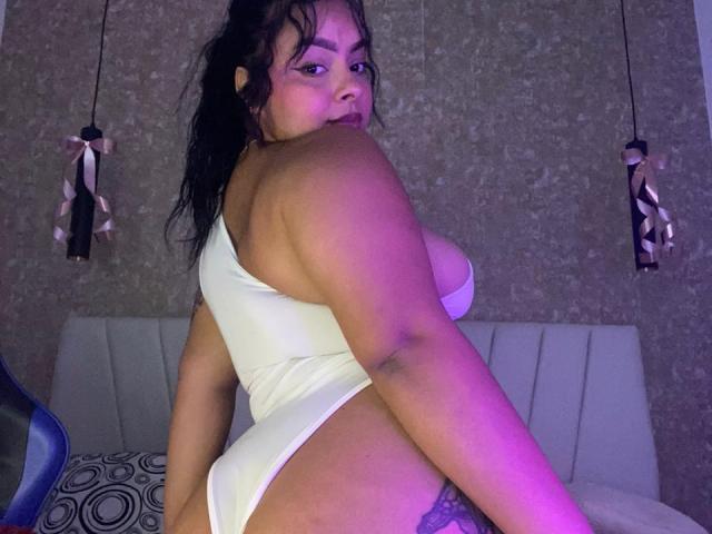 AmarantaCarey with dark hair, medium skin, and full tits in a white one-piece, looking back, showing her ass and a tattoo on her right thigh. Purple light.
