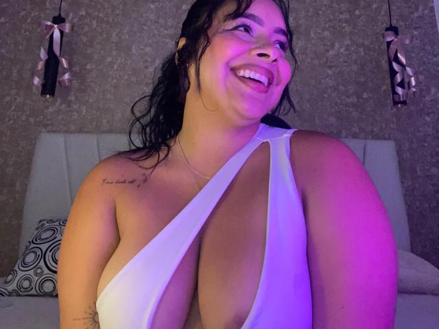 AmarantaCarey with large tits in a white, deep-cut, revealing top, smiling, pink and purple lighting, black wavy hair, tattoo on right shoulder, and a necklace.