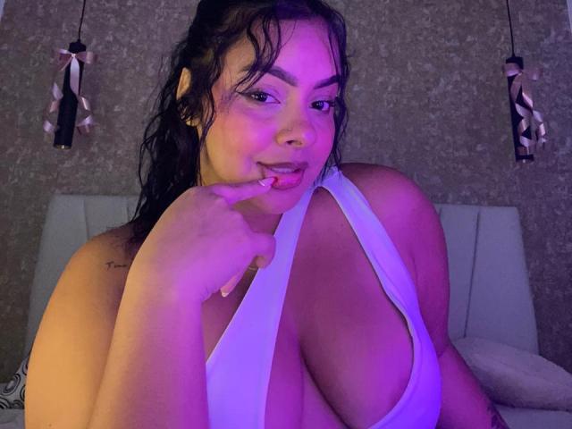 AmarantaCarey with wet black hair, wearing a white halter top that reveals large, round tits, biting her finger, looking at the camera with purple lighting.