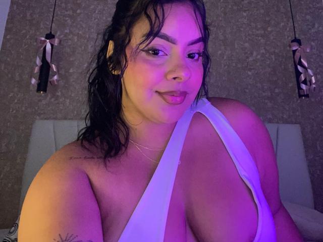 AmarantaCarey with wet black hair, wearing a low-cut white top, showing large boobs, purple-lit, smiling, tattoo on left shoulder, earrings, and a necklace.