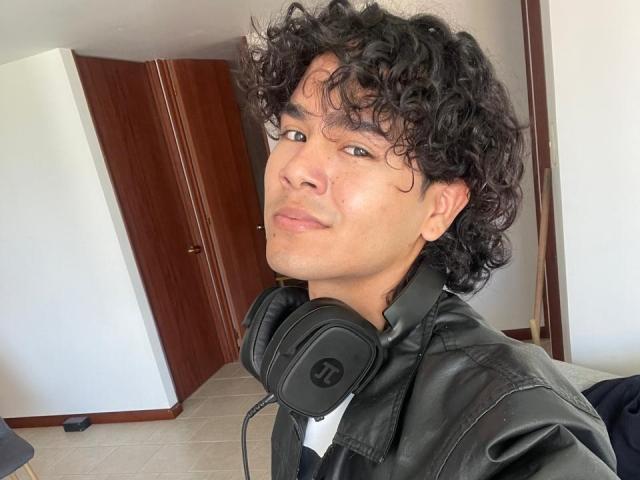 AndruStar with curly black hair, light brown skin, and a slight smirk, wearing a black leather jacket and black headphones, looking at the camera. AndruStar's nose is slightly shiny.