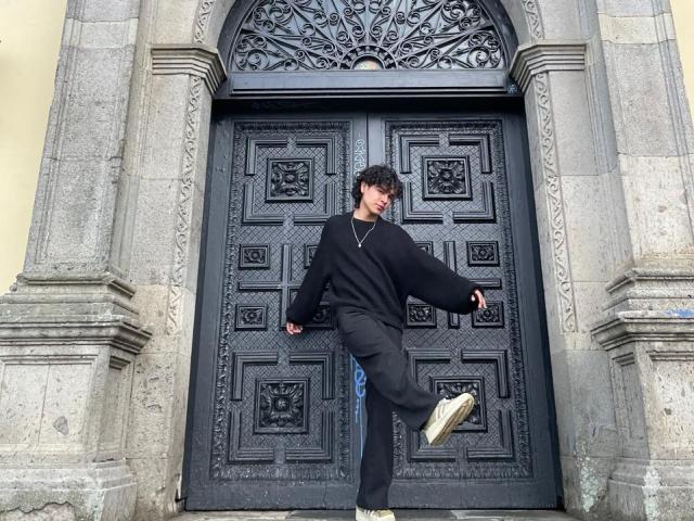 AndruStar is a dark-skinned dude with curly hair, wearing a black sweater and pants, standing in front of ornate black doors, mid-step with one foot up, smiling.