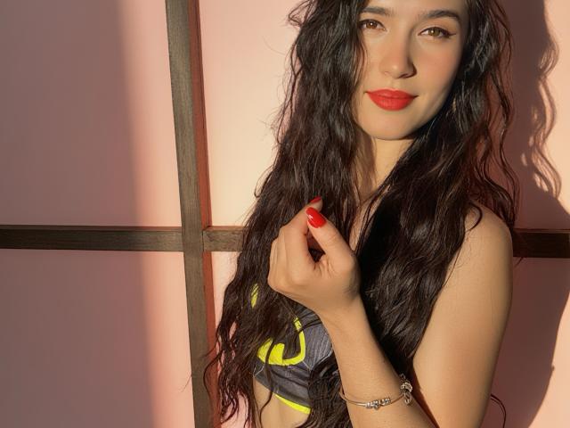ChlooeZoe with long wavy black hair, red lipstick, and red nail polish, holding her hair, wearing a gray sports bra with yellow accents, sunlight casting shadows.