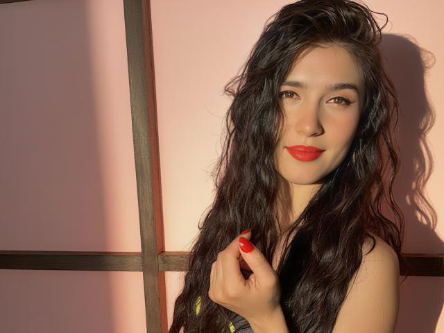 ChlooeZoe with long, wavy black hair, red lipstick, and red nails, holding her hair, smiling, brown eyes, fair skin, wearing a black strap, sunlight on her face.