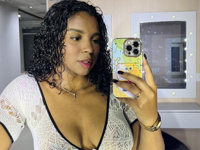 LisaMendez with curly black hair, medium brown skin, wearing a white lace top, showing her large tits, holding a phone with a yellow case, pink lipstick, black nails.