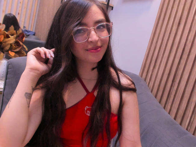 EvaMcAllister with long dark hair, glasses, red halter top, small tits, tattoo on right arm, smiling, holding hair, sitting on gray couch, wooden wall in background.