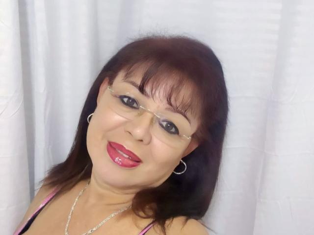 HornyFemme69 has brown hair, glasses, red lipstick, and is smiling with shiny pink lips. She's wearing a pink top and silver hoop earrings, against a white curtain.