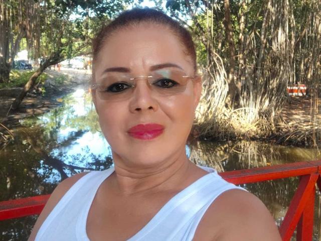 HornyFemme69 with medium brown skin, red lipstick, and white tank top, wearing glasses, standing by a red railing, next to a reflective waterway, in a sunlit wooded area.