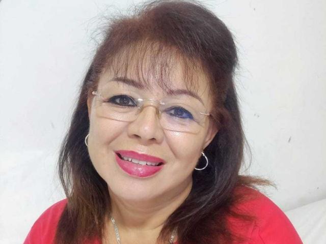 HornyFemme69 has brown hair, glasses, pink lipstick, red shirt, and silver hoop earrings, smiling with blue eye makeup, showing her shiny teeth.