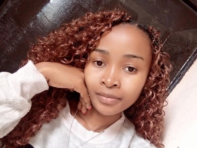 Samyra69 with curly brown hair, brown eyes, and light brown skin, wearing a white sweater, resting her head on her hand, smiling slightly, showing no makeup.