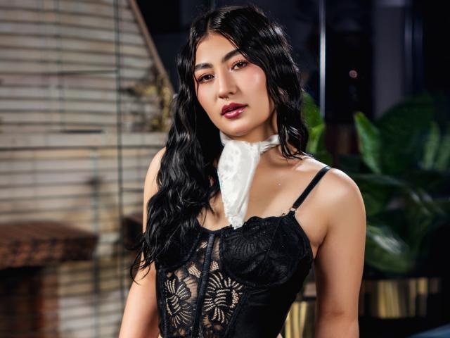 MaddisonCarter with long black wavy hair, wearing a black lace corset, white scarf, and dark red lipstick, standing in a dimly lit room, showcasing her tits.
