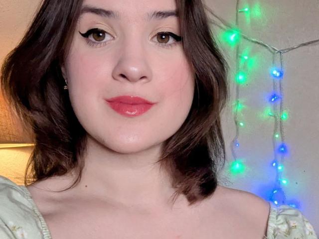 ClickWoman has shiny red lips, brown wavy hair, and black eyeliner. She's wearing a light green top with floral patterns. Her cute ass is barely visible.
