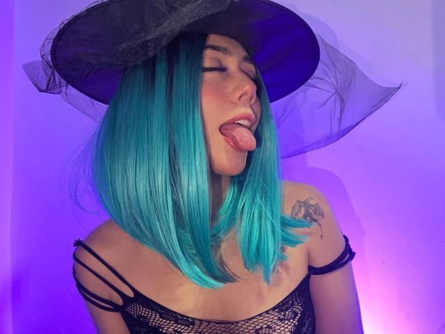 AlannaJonson with teal hair, black witch hat, tongue out, black mesh top, tattoo on left shoulder, purple-blue lighting. Boobs visible through mesh. AlannaJonson's tongue is out.