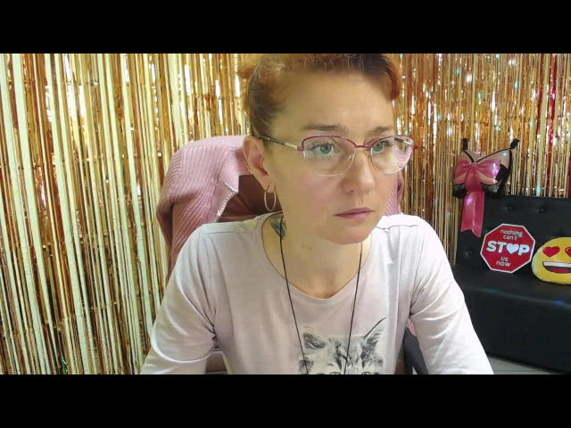 AmeliRox with red hair, glasses, pale skin, and a white shirt, sitting in a chair, looking slightly confused, wearing a necklace, and a pink cloth behind her.