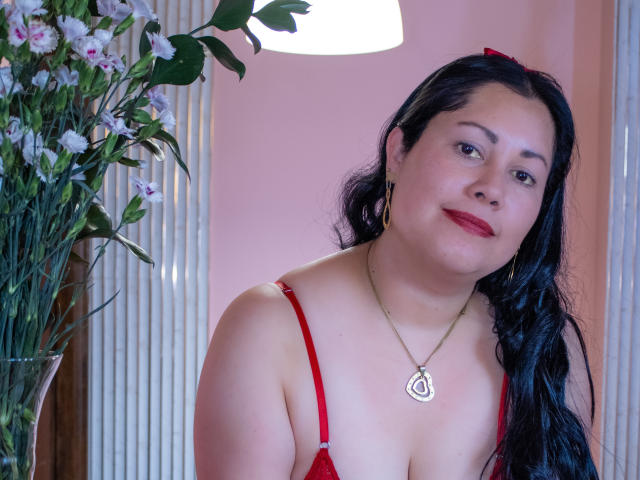 MarianaSmitt, avec de longs cheveux noirs, un soutien-gorge rouge, un collier et du rouge à lèvres rouge, souriant à la caméra, un nœud rouge dans les cheveux, des fleurs sur la gauche, une lampe blanche au-dessus, un mur rose en arrière-plan..