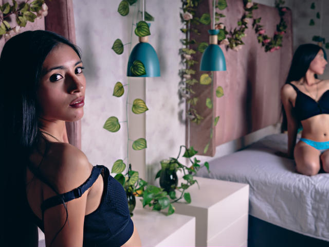 AlanaMoore à quatre pattes, portant un soutien-gorge noir et des culottes en dentelle bleue, avec de longs cheveux noirs, regardant la caméra, montrant ses fesses et ses petites seins. Décor d'une chambre..