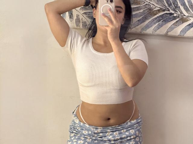 AmeliaWilson has brown hair, a light pink shirt, and a slight smile. She's showing a small nipple on her shirt. Shelves with books are in the background.
