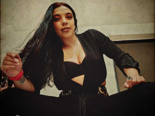 Mavis69 with long black hair, wearing a black zip-up top and bra, showing her tits, black pants, red bracelet, and tattoo on right wrist, sitting confidently.