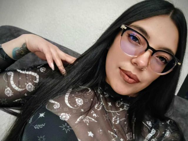 Mavis69 with long black hair, glasses, nose ring, and lip ring, wearing a sheer black top with moon and star patterns, showing a tattoo on her right hand.