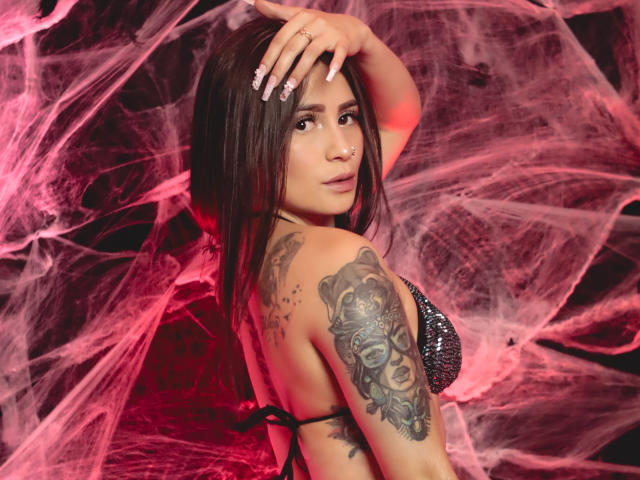 NinaBellaG with long brown hair, looking back, wearing a sparkly black bra, hand on head, showing a detailed skull tattoo on her right arm, red and pink smoke.