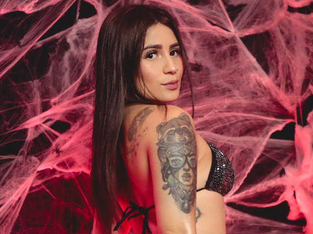 NinaBellaG with long brown hair, wearing a sparkly black bikini top, looking back with a nose ring. She's got a detailed tattoo on her right arm, showing a face.