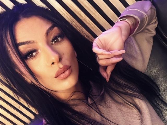 KinkyAmelie with long black hair, pouty lips, and brown eyes, wearing a beige top, right hand resting on a black leather surface, fingers slightly curled.