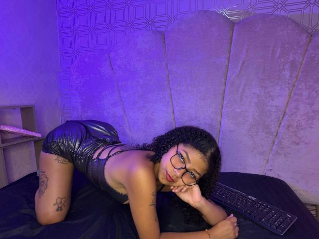 MistresSamanta, black curly hair, glasses, black leather booty shorts, on all fours on a bed, purple light, tattoos on thigh and arm, black keyboard in front.