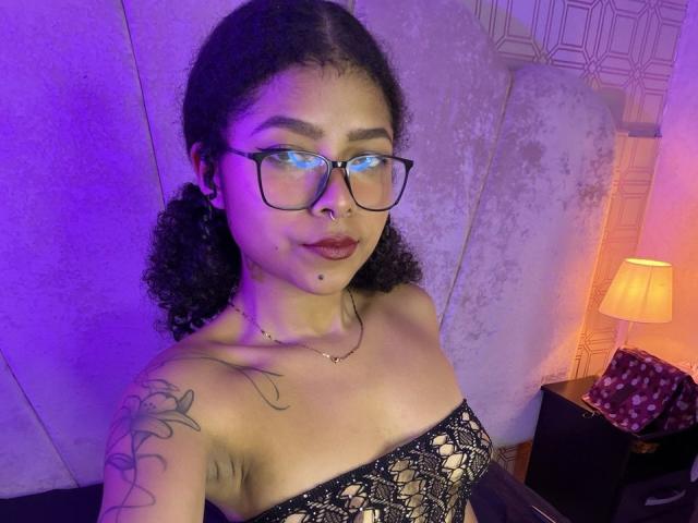 MistresSamanta with curly black hair, glasses, nose ring, and lipstick, wearing a lace strapless top, showing a tattoo on her right shoulder, in purple and orange lighting.