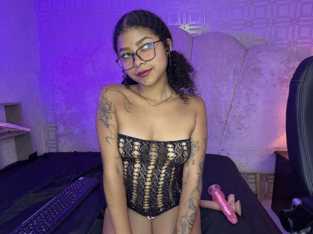 MistresSamanta with curly black hair, glasses, and tattoos, wearing a black lace-up bustier, holding a pink dildo, sitting on a bed, under purple lighting.