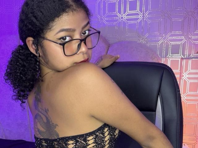 MistresSamanta with curly black hair, glasses, and a tattoo on her back, looking over her shoulder at the camera. She's wearing a black, netted dress, showing her bare back.