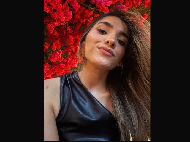 AdharaCooperr with long brown hair, wearing a black leather sleeveless top, smiling at the camera, standing in front of vibrant red flowers, looking sexy and confident.