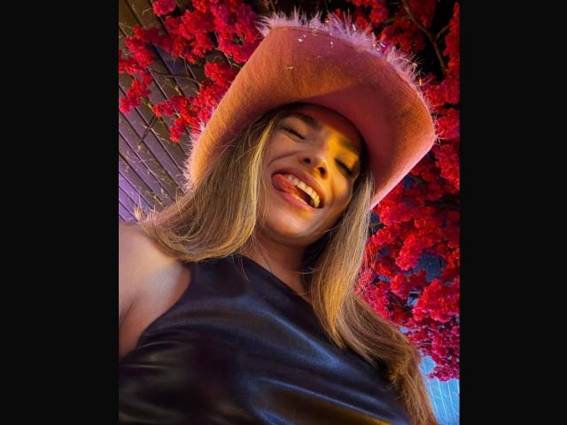AdharaCooperr with long blonde hair, sticking out tongue, wearing pink fuzzy hat and black leather top, red flowers in background, low-angle shot, showing her face and upper body.