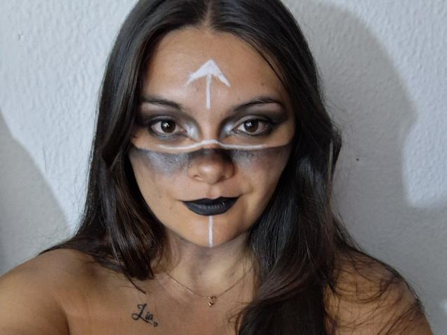 KatherinLatin with dark makeup, white face paint lines, black lipstick, and tattoo on her chest, standing against a white textured wall, showing off her face and upper body.