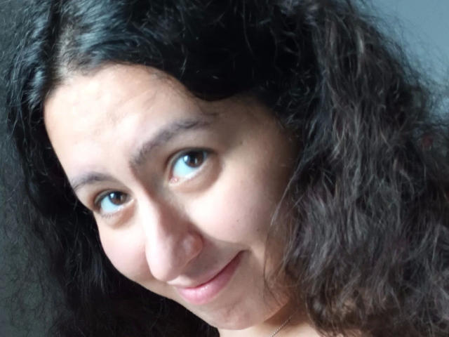 El rostro de Linasardi-hot, mostrando cabello negro rizado, ojos marrones y una ligera sonrisa, con una nariz pequeña y labios carnosos, en una luz suave y natural..