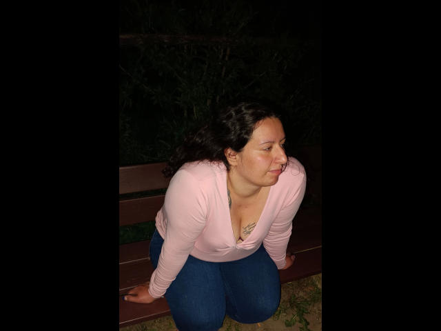 Linasardi, avec ses cheveux noirs bouclés, son sweat à capuche rose et son jean bleu, est accroupie sur un banc, regardant sur le côté avec une légère moue, portant un collier, la nuit..