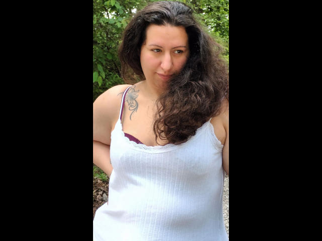 Linasardi-hot tem cabelos castanhos cacheados, uma tatuagem no ombro direito e veste uma regata branca que mostra seu sutiã roxo. Suas mãos estão atrás das costas..