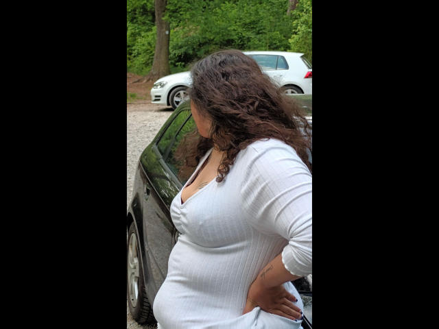 Linasardi-hot with curly brown hair, wearing a tight white ribbed dress, showing her big ass and large tits, standing next to a black car in a gravel area.