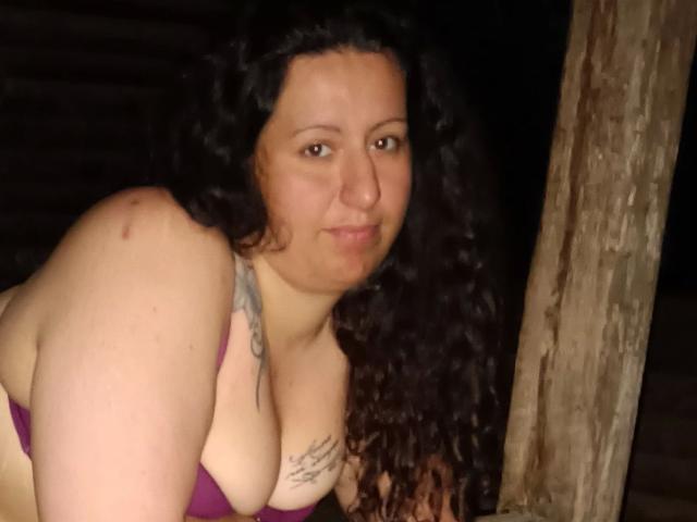 Linasardi-hot heeft krullend zwart haar, een lichte huid, kleine tatoeages en draagt een paarse beha, waardoor haar grote borsten zichtbaar zijn, evenals een lichte blauwe plek op haar rechter schouder..