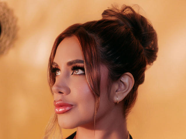 SarayYorks has shiny red lips, subtle makeup, and a messy bun with loose brown hair. Her green eyes are slightly upturned, and she's wearing small hoop earrings.